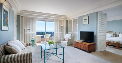 Image of a Superior Suite living room with pastel-toned interiors, a sofa for three, a table, and a television overlooking the sea.